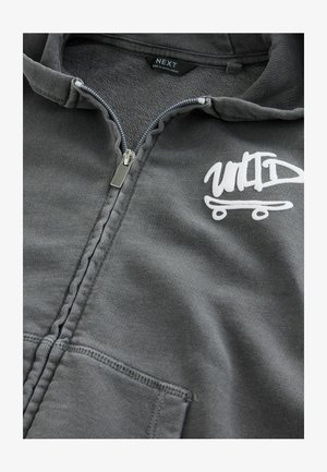 Pilka užtrauktuku džemperis, pagamintas iš švelnios medžiagos. Turi priekinę kišenę, sidabrinį užtrauktuką ir baltą grafikos logotipą su riedlenčių dizainu.