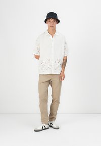 Hvid kortærmet skjorte med blomsterudskæringer, parret med beige bukser og sort bucket hat. Sneakers har hvide og grå accenter.