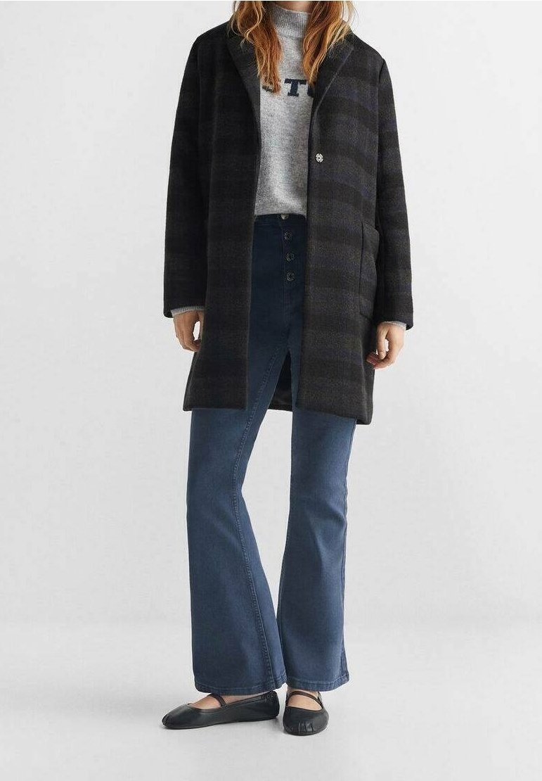 Cappotto a quadri nero e grigio con chiusura a bottone, indossato sopra un maglione grigio e pantaloni a zampa d'elefante blu scuro. Scarpe basse nere decorate con ritagli.