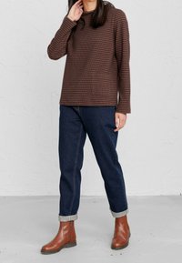 Osoba v hnědém pruhovaném dlouhém tričku, tmavě modrých džínech s manžetami a hnědých kožených kotníkových botách stojí proti bílé strukturované zdi.