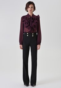 Paarse doorzichtige blouse met ruches bij de halslijn, gecombineerd met zwarte, hooggetailleerde broek met gouden knoopaccenten.