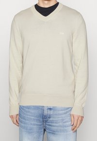 Beżowy sweter w serek z miękkiego dzianiny z długimi rękawami, z prążkowanymi mankietami i dolnym brzegiem; małe logo na piersi; noszony na czarnej koszulce.