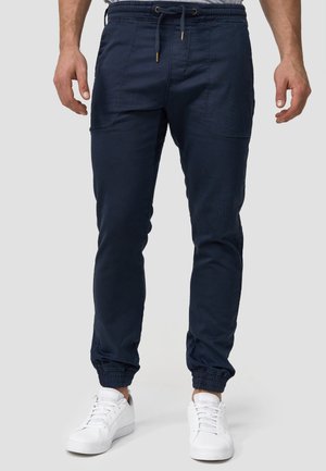 Navy Jogginghosen aus leichtem Stoff, ausgestattet mit einem elastischen Bund, einer Kordel, Seitentaschen und elastischen Bündchen. Dazu weiße Sneakers.
