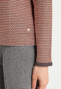 Dzianinowy sweter z wzorem w kolorach pomarańczowym, białym i granatowym; ściągacze przy mankietach; w połączeniu z szarą tkaniną o wzorze jodełki. Widoczny złoty akcent.