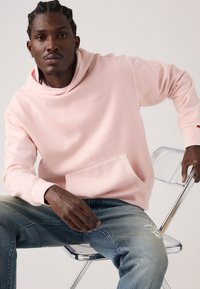 Ανοιχτό ροζ φούτερ με κουκούλα από απαλό ύφασμα, με τσέπη μπροστά και άνετη εφαρμογή. Συνδυασμένο με σκισμένο μπλε τζιν και διαφανή καρέκλα.