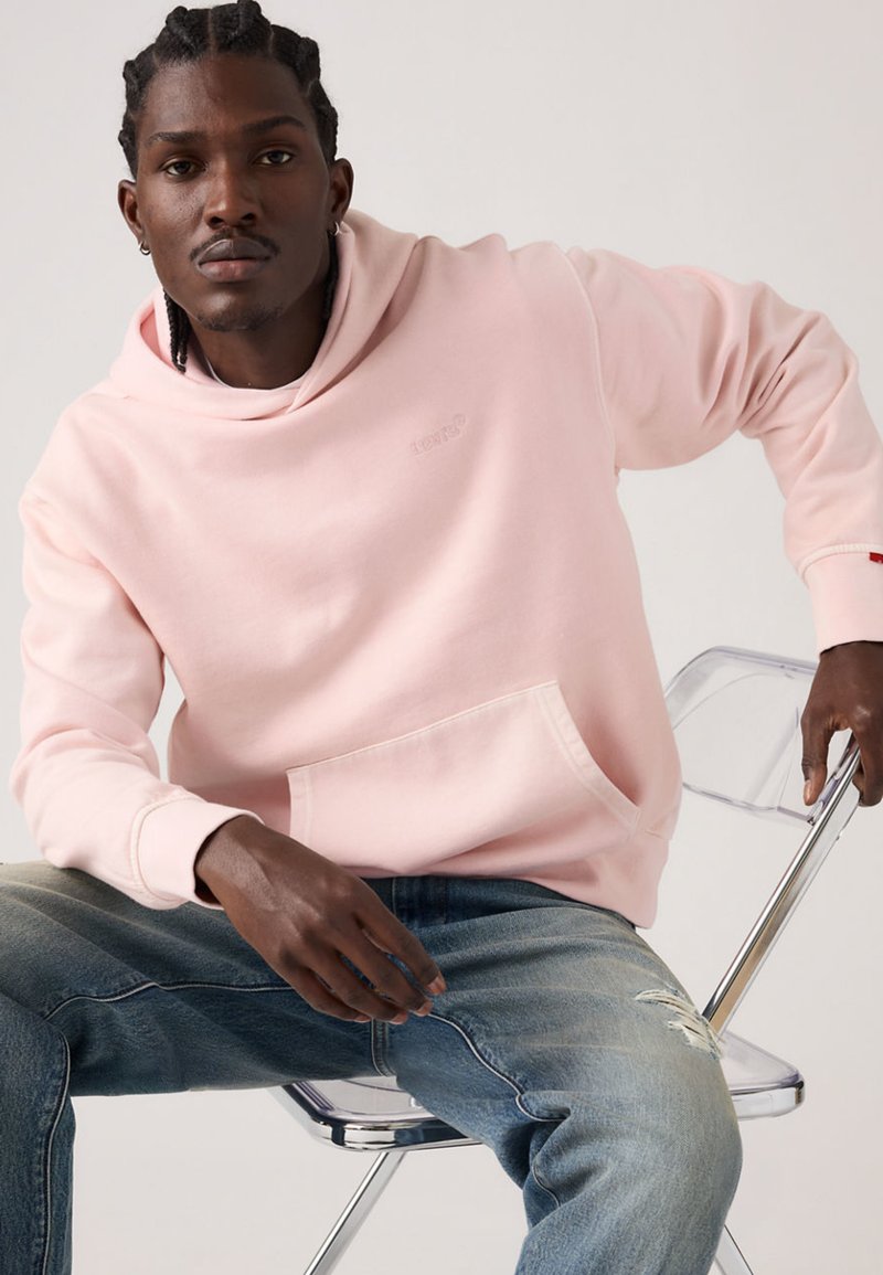 Sweat à capuche rose clair en tissu doux, avec une poche avant et une coupe décontractée. Associé à un jean bleu déchiré et une chaise transparente.