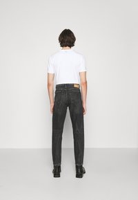 Persona con cabello corto castaño, vistiendo una camiseta blanca, jeans negros y botas negras, de pie frente a una pared blanca sencilla.