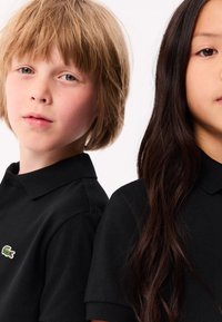 En blond pojke och en mörkhårig flicka står rygg mot rygg, båda iförda svarta polo-tröjor med en liten grön krokodil-logotyp.