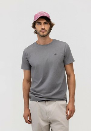 Hombre con una camiseta gris, pantalones beige claro y una gorra de béisbol rosa descolorida, de pie frente a un fondo claro y liso.