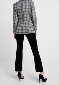 Žena v černých rozšiřujících kalhotách a černých vysokých podpatcích, oblečená v šedooranžovém kostkovaném saku, stojí na jednolitém pozadí.