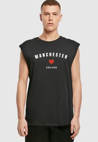 Mustavärinen hihaton t-paita, jossa on valkoisella painettuna "MANCHESTER", punaisen sydämen grafiikka ja "ENGLAND" valkoisena alla. Pehmeää kangasta, rento malli.