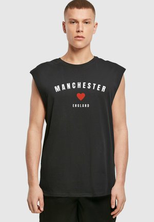Sort ærmeløs t-shirt med "MANCHESTER" trykt i hvidt, et rødt hjertegrafik, og "ENGLAND" i hvidt nedenunder. Blødt stof, afslappet pasform.