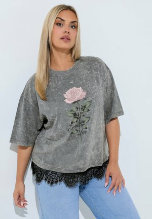 Vrouw met lang blond haar die een grijs oversized T-shirt draagt met een roze rozenprint en zwarte kanten zoom, gecombineerd met lichtblauwe spijkerbroek.