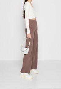 Weißes geripptes Langarmoberteil, braune plissierte weit geschnittene Hose, weiße Sneaker mit gelben Akzenten und eine kleine handliche Handtasche mit blauem Muster.