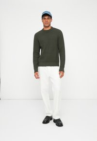 Maglione a maglia texture verde oliva con colletto rotondo, abbinato a pantaloni bianchi a gamba dritta e scarpe nere con lacci. Cappellino di denim blu indossato.