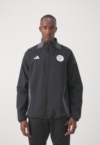 Chaqueta deportiva negra con detalles grises, cierre completo, cuello alto y parche con logotipo. Tejido ligero y puños elásticos para un diseño ajustado.