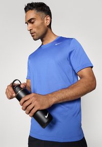 Blå atletisk T-shirt med korte ærmer og rund hals, parret med en sort isoleret vandflaske med en sølvring og håndtag.
