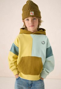 Kind met een groene gebreide muts en een hoodie in kleurblokken met gele, blauwe en bruine delen, staand met handen in de zakken tegen een effen achtergrond.