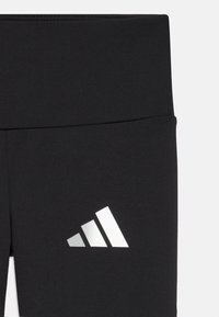 Svarta leggings med en slät textur, med en silvertriangulär logotyp på vänster sida och ett elastiskt midjeband högst upp.