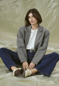 Šedý houndstooth blazer přes bílý tričko, doplněný tmavými širokými jeansy a béžovými botami. Sedící pose na texturovaném povrchu.