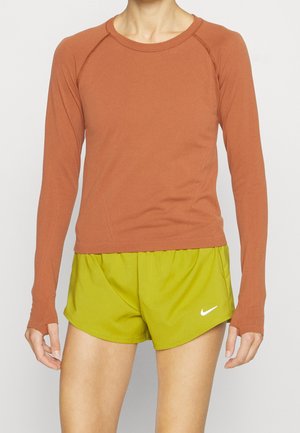 Rdzawy top z długim rękawem i otworami na kciuki, w połączeniu z jasnymi zielonymi szortami z małym białym logo Nike. Miękki, elastyczny materiał.