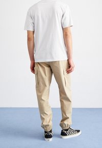 Pantalons cargo beiges avec des poignets élastiques, poches latérales, portés avec un t-shirt blanc uni et des baskets à carreaux noirs, sur un fond simple.