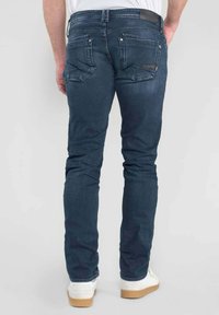Jean slim fit bleu foncé avec une finition texturée, comprenant deux poches arrière et une étiquette en cuir marron à la taille.