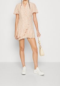 Robe portefeuille fleurie pêche avec manches courtes volantées, ourlet à volants, associée à des baskets blanches et un petit sac à main crème.