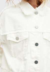 Giacca di denim bianco con dettagli sfrangiati, chiusure con bottoni e due tasche sul petto. Presenta un collo classico e tessuto strutturato.