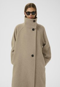 Cappotto beige simile alla lana con colletto alto, dotato di chiusura frontale con bottoni neri. Vestibilità lunga e comoda con maniche raglan.