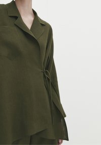 Camicia in lino leggera di colore verde oliva, con colletto a pattina, legatura laterale e vestibilità ampia. Presenta una tasca sul petto e orlo asimmetrico.