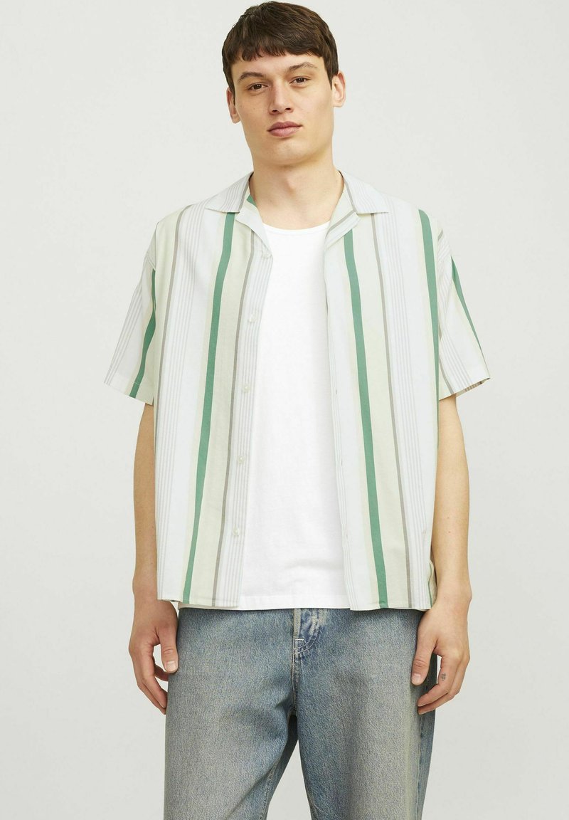 Camicia a maniche corte in tessuto leggero con strisce verticali verdi e bianche, design con bottoni sul davanti e vestibilità rilassata. Indossata sopra una maglietta bianca.