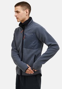 Man in gray fleece jacket with zippered chest and side pockets, standing in profile with hands clasped at waist.