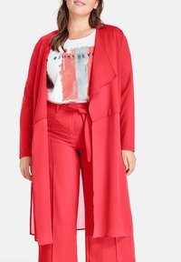 Mujer vistiendo una camiseta gráfica blanca, pantalones rojos de pierna ancha con un lazo en la cintura, y un cárdigan largo rojo abierto, contra un fondo liso.