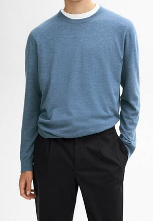 Mand iført en lyseblå langærmet sweater over en hvid undertrøje og sorte bukser, stående med den ene hånd i lommen.