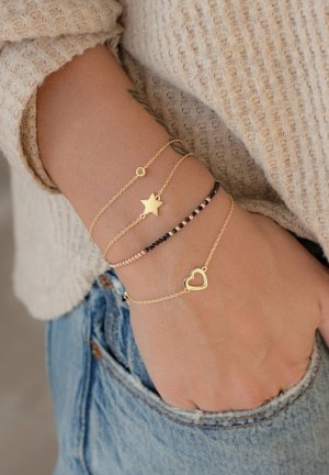 Goldene Kettenarmbänder mit einem Stern- und einem Herzanhänger, abwechselnd mit schwarzen und rosafarbenen Perlen, getragen an einem Handgelenk mit Jeans.