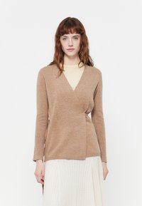 Beige, puha anyagú kötött pulóver, hosszú ujj�ú és mély V-nyakkal. Világos színű blúz fölött viselve, egy textúrált fehér szoknyával párosítva.