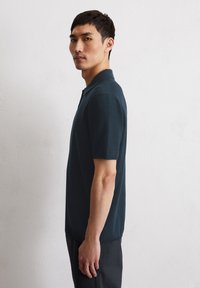 Ciemnoniebieska koszulka polo z krótkimi rękawami, o teksturowanej tkaninie, klasycznym kołnierzyku i prostym kroju, noszona przez modela zwróconego bokiem.