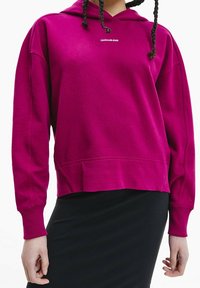 Fuchsia sweatshirt med huva och ribbade ärmslut, med en liten vit "Calvin Klein Jeans"-logotyp på bröstet, kombinerad med en svart kjol.