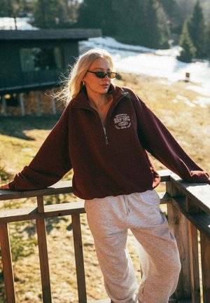 Blondynka w okularach przeciwsłonecznych, bordowej bluzie i szarych spodniach dresowych opiera się o drewnianą barierkę balkonu w słonecznym, zaśnieżonym górskim otoczeniu.