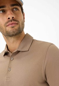 Polo shirt color sabbia con colletto, due bottoni frontali e tessuto strutturato, mostrato su un modello dalla spalla in su.