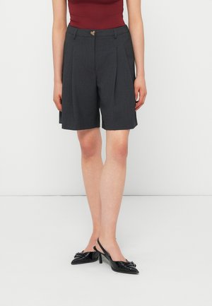 Femme portant un short plissé gris foncé, un débardeur bordeaux, et des escarpins slingback noirs à bout pointu, debout sur un sol blanc.