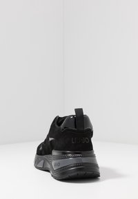 Sneaker nera Liu Jo con suola spessa e texture, dettagli in suede e lucidi, e linguetta sul tallone, posizionata su una superficie bianca con sfondo neutro.