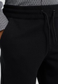 Svarta vävda joggers med elastisk midja, snörning och texturerat tyg. Har sidofickor och subtila korshatch-detaljer.