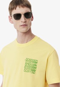 T-shirt en coton jaune avec un col rond et des manches courtes, présentant un imprimé graphique vert du mot "Lacoste" sur la poitrine.