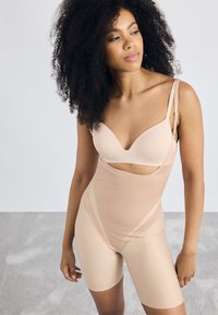 Beige bodysuit, jossa on mukautuva muotoilu, läpikuultavia paneeleja ja säädettävät hihnat, pituus keskikohtaan. Silkkinen, joustava kangas.