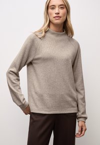 Pull en tricot beige avec un col haut, de longues manches raglan, des poignets et un ourlet côtelés. Porté sur un pantalon marron foncé, mettant en avant une coupe décontractée.