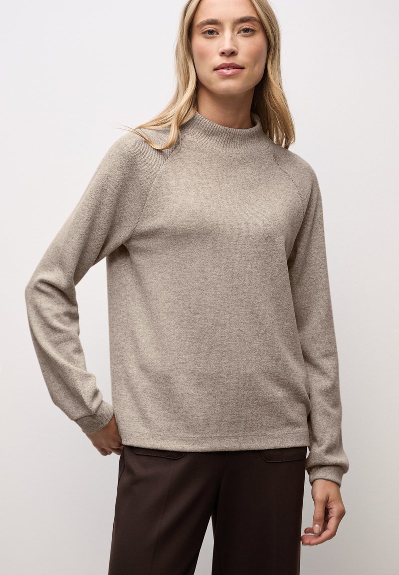 Pull en tricot beige avec un col haut, de longues manches raglan, des poignets et un ourlet côtelés. Porté sur un pantalon marron foncé, mettant en avant une coupe décontractée.