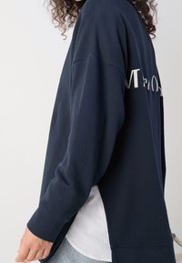 Sudadera de gran tamaño en color azul marino con un diseño impreso en blanco en la parte posterior, que cuenta con un dobladillo redondeado y mangas de corte holgado.