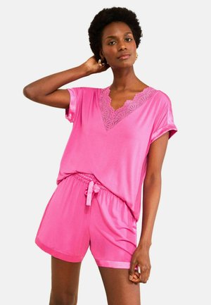 Woman with short curly hair wears bright pink lace-trimmed V-neck top and matching drawstring shorts, posing with one hand behind her head.